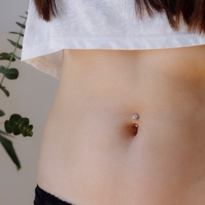 Puede incluir: Primer plano del abdomen de una persona con un piercing en el ombligo. El piercing es una barra de plata con una pequeña gema transparente en forma de flor en la parte superior.