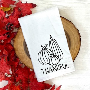 Puede incluir: Toalla de cocina blanca con un contorno negro de tres calabazas y la palabra "THANKFUL" impresa debajo.