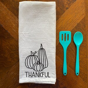 Puede incluir: Toalla de cocina blanca con un contorno negro de tres calabazas y la palabra "THANKFUL" impresa debajo. La toalla est&aacute; sobre una mesa de madera con dos utensilios de cocina de color turquesa.