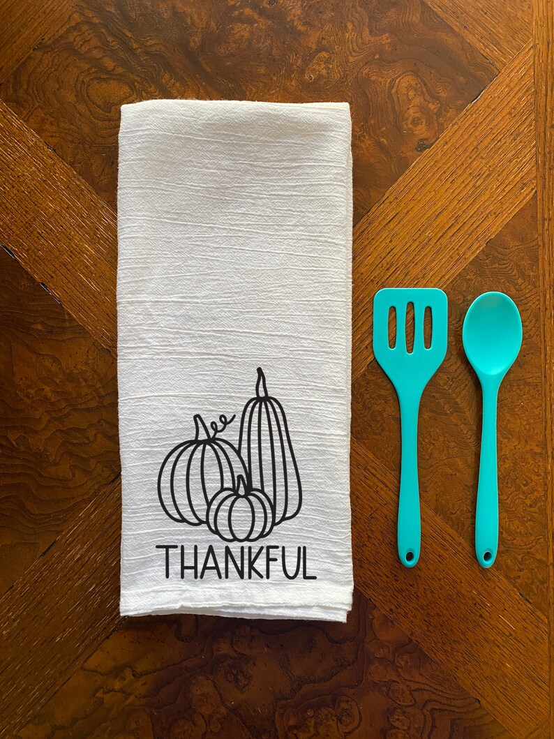 Puede incluir: Toalla de cocina blanca con un contorno negro de tres calabazas y la palabra "THANKFUL" impresa debajo. La toalla est&aacute; sobre una mesa de madera con dos utensilios de cocina de color turquesa.