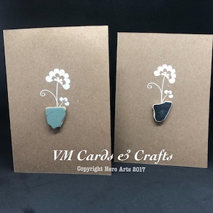 May include: Two handmade greeting cards featuring a white flower design and a small pot made from a broken piece of tile. The pot on the left card is light blue and the pot on the right card is black. The cards are on a brown background and have the text "VM Cards & Crafts Copyright Hero Arts 2017".