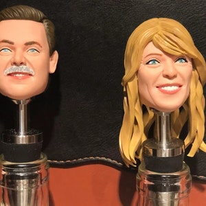 May include: Two bottle stoppers with realistic faces. The bottle stopper on the left features a man with a mustache and brown hair. The bottle stopper on the right features a woman with blonde hair.