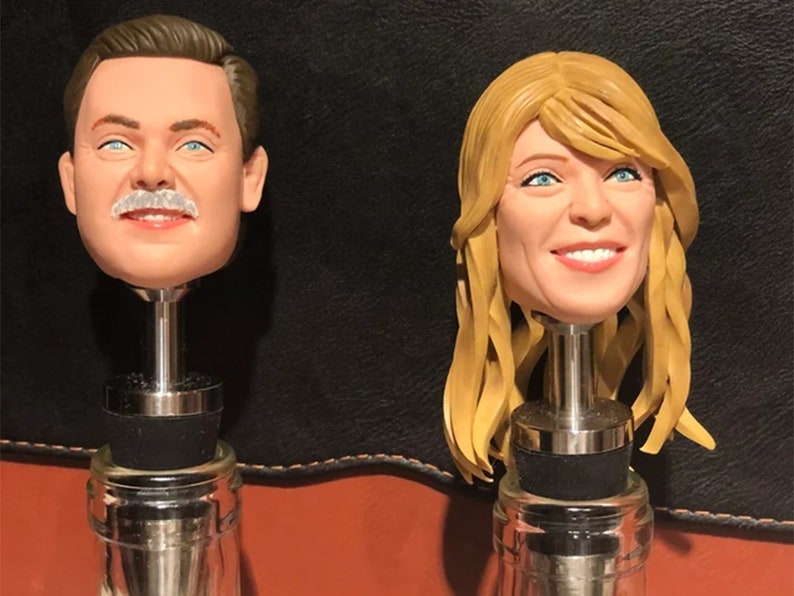 May include: Two bottle stoppers with realistic faces. The bottle stopper on the left features a man with a mustache and brown hair. The bottle stopper on the right features a woman with blonde hair.