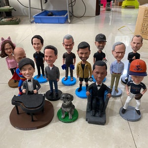 May include: A collection of 12 custom bobbleheads, including a baseball player, a musician playing a piano, a dog, and several people in various poses. The bobbleheads are displayed on a table with personalized bases.