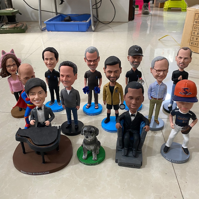 May include: A collection of 12 custom bobbleheads, including a baseball player, a musician playing a piano, a dog, and several people in various poses. The bobbleheads are displayed on a table with personalized bases.