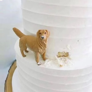 May include: A golden retriever cake topper figurine stands on a tiered white cake. The dog is light brown with a detailed fur texture. The cake has a textured, ridged design.