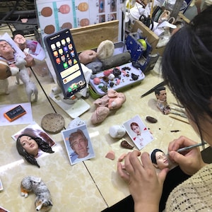 May include: A person is working on a clay sculpture of a person's face. The sculpture is on a table with other tools and materials, including a phone, a small dog sculpture, and a photo of a person. The person is using a small tool to shape the clay.