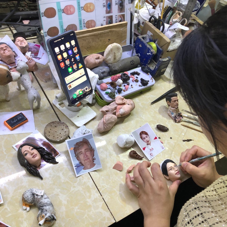 May include: A person is working on a clay sculpture of a person's face. The sculpture is on a table with other tools and materials, including a phone, a small dog sculpture, and a photo of a person. The person is using a small tool to shape the clay.
