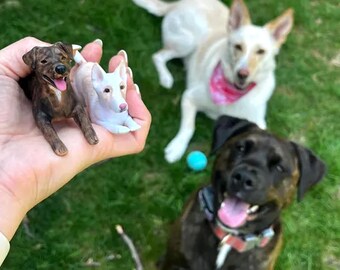 May include: A hand holding two small dog figurines, one brown and one white, with a real dog in the background. The brown figurine has its tongue out. Two other dogs are in the background, one wearing a pink bandana.