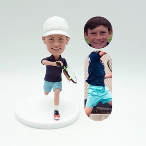 May include: A custom bobblehead figurine of a person playing tennis. The figurine is wearing a white hat, a blue shirt, and blue shorts. The person is holding a tennis racket and is in a running pose. The figurine is on a white base.