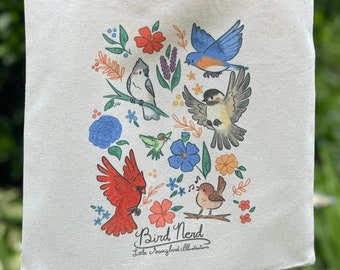 May include: A white canvas tote bag with a colorful design of birds and flowers. The text "Bird Nerd" and "Little Fromgbird Illustrations" is printed on the bag.
