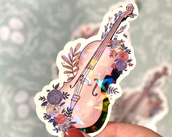 May include: A holographic cello sticker with floral details. The cello is decorated with pink, red, blue, and yellow flowers. The sticker is on a white background.