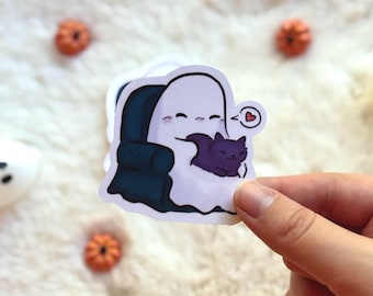 Op de afbeelding: Een sticker met een cartoongeest die in een donkerblauwe fauteuil zit en een paarse kat knuffelt. Een klein rood hart zweeft boven het hoofd van de geest. De sticker wordt door een hand vastgehouden, met kleine oranje pompoenversieringen op de achtergrond.