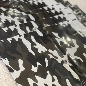 Könnte beinhalten: Ein Stapel Blätter mit Tarnmuster. Die Blätter weisen ein sich wiederholendes Muster aus weißen, schwarzen und braunen Formen auf. Die Blätter sind glänzend und scheinen aus einem flexiblen Material zu bestehen.