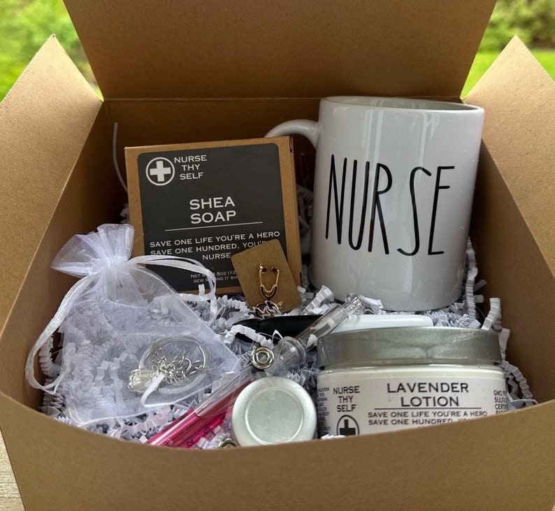 May include: A brown cardboard box filled with a gift set for a nurse. The box contains a white mug with the word "NURSE" in black lettering, a white jar of lavender lotion, a white bar of shea soap, a small white bag with a stethoscope charm, and a pink lip balm.
