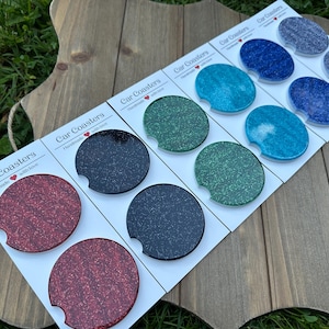 May include: A collection of car coasters in various colors, including red, black, green, blue, and gray, with glittery finishes. The coasters are arranged on a white card with the text "Car Coasters Handmade with love".