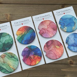 May include: Set of six car coasters with abstract watercolor designs in shades of blue, green, pink, and orange. Each coaster features a unique design with gold accents. The coasters are labeled "Car Coasters Handmade with love".