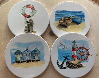 May include: Set of four white ceramic coasters with different beach-themed designs. One coaster features a red and white life preserver with two seagulls perched on a wooden post. Another coaster shows two boats on a sandy beach with blue water and white waves. A third coaster has three blue beach huts on a sandy beach with a blue sky. The final coaster has a red and white lighthouse with a ship's wheel and an anchor on a rocky shore.