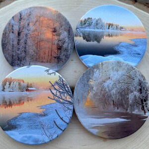 Peut inclure: Ensemble de quatre sous-verres ronds représentant des scènes d'hiver avec des arbres givrés et un lac reflétant un lever de soleil. Les sous-verres sont en céramique et ont une finition brillante.