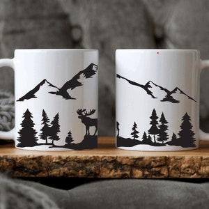 Può includere: Due tazze in ceramica bianca con silhouette nere di montagne e foreste. Una tazza presenta una silhouette di alce.