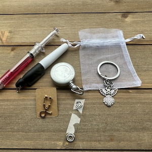 May include: A set of medical themed gifts including a retractable badge reel, a stethoscope charm, a syringe pen, a white marker, and a silver angel keychain in a white organza bag.