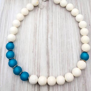 May include: A necklace with white and teal beads. The necklace has large, round, textured white beads alternating with teal beads. It is strung on a silver chain and has a silver clasp.
