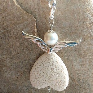 Könnte beinhalten: Ein weißer Lavastein-Engel-Anhänger mit einem silbernen Metall-Heiligenschein und Flügeln. Der Engel hat eine weiße Perle als Kopf.