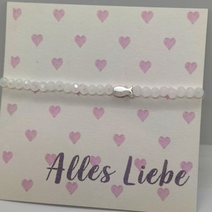 Könnte beinhalten: Ein weißes Perlenarmband mit einem silbernen Fischanhänger. Das Armband wird auf einer Karte mit einem Muster aus rosa Herzen und dem Text "Alles Liebe" in lila Schrift präsentiert.