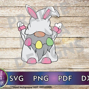 Puede incluir: Un gnomo blanco que lleva un disfraz de conejito rosa y blanco con orejas rosas y una capucha blanca. El gnomo tiene una barba gris con tres huevos de Pascua coloridos colgando.