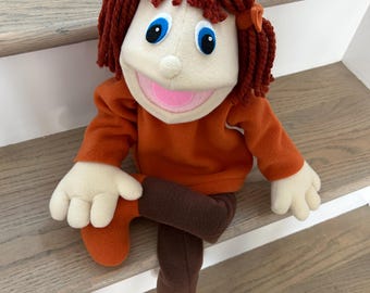 May include: A hand puppet with a cream-colored face, blue eyes, and a pink mouth. The puppet has dark red yarn hair styled with two small orange bows. It wears an orange long-sleeved shirt, dark brown pants, and orange shoes made of soft fabric.
