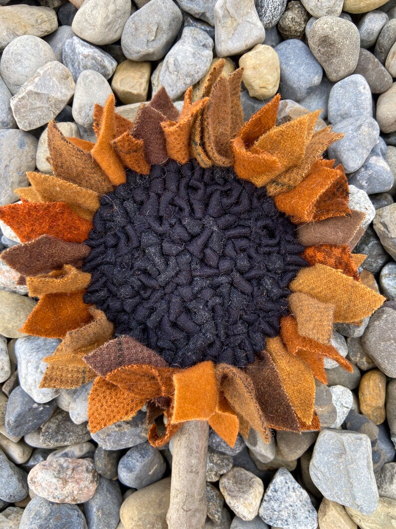 May include: A handmade felt sunflower with brown, orange, and black petals. The sunflower is attached to a wooden stick.