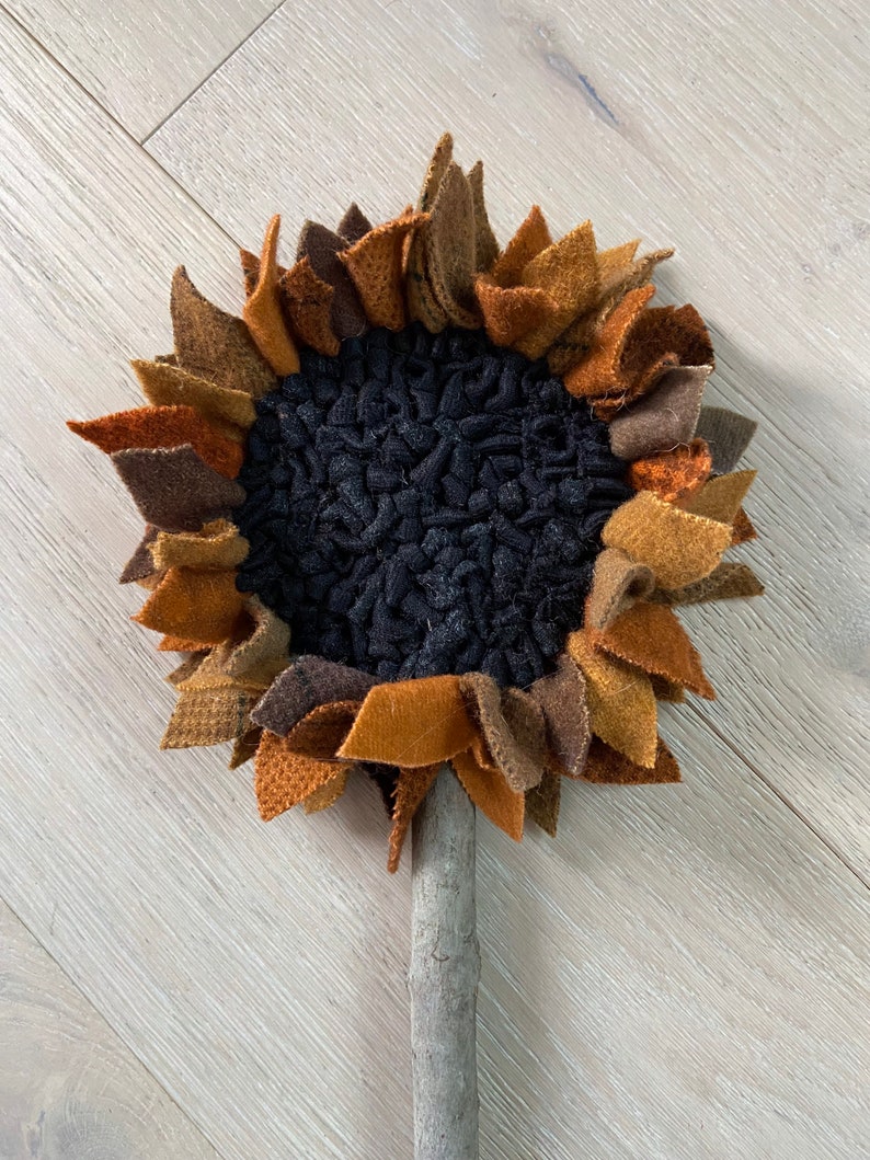 May include: A handmade felt sunflower with brown, orange, and black petals. The sunflower is attached to a wooden stick.