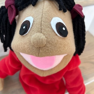 May include: A handmade puppet with a tan face, black yarn hair styled in two pigtails with burgundy bows, and a red hoodie. The puppet has large, expressive eyes and a pink tongue.