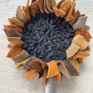 May include: A handmade felt sunflower with a brown stem. The sunflower has a black felt center and petals in shades of brown, orange, and yellow.