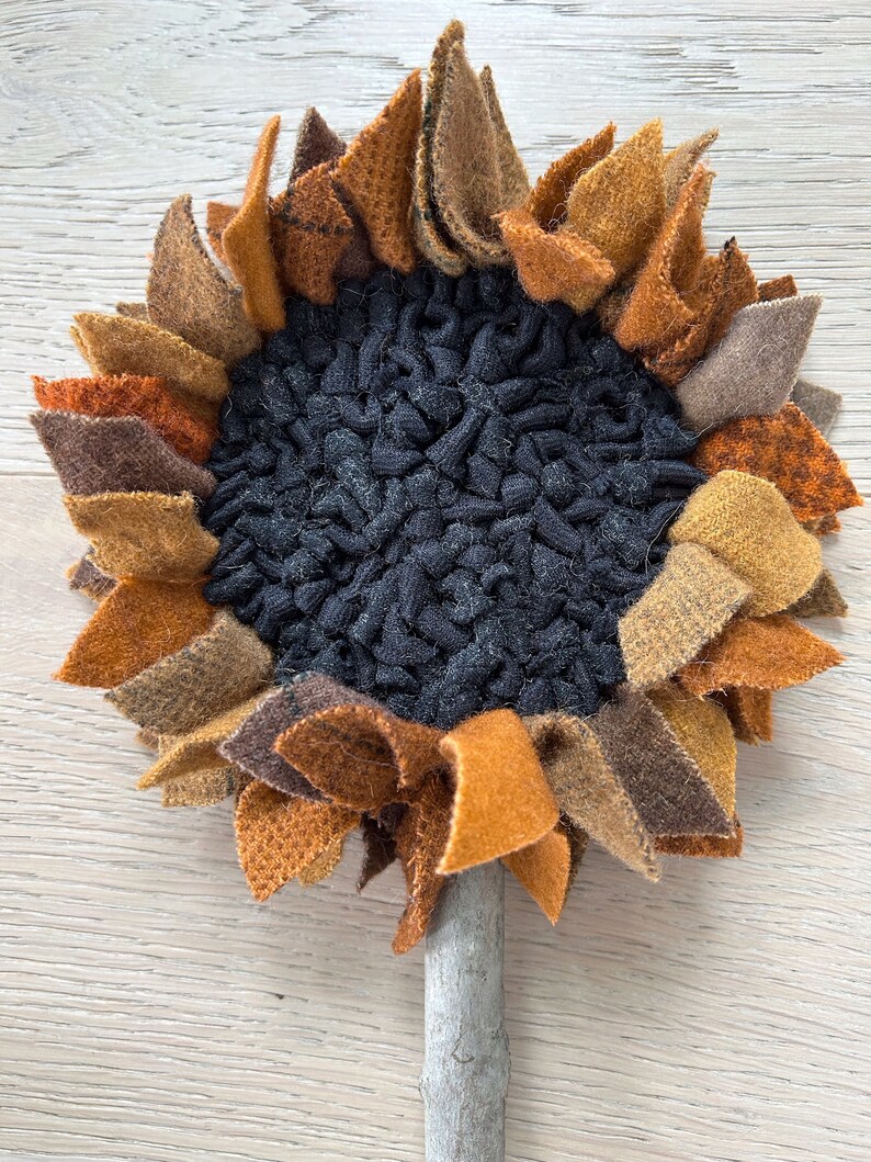May include: A handmade felt sunflower with a brown stem. The sunflower has a black felt center and petals in shades of brown, orange, and yellow.