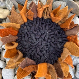 May include: A handmade felt sunflower with brown, orange, and black petals. The sunflower is sitting on a bed of gray rocks.