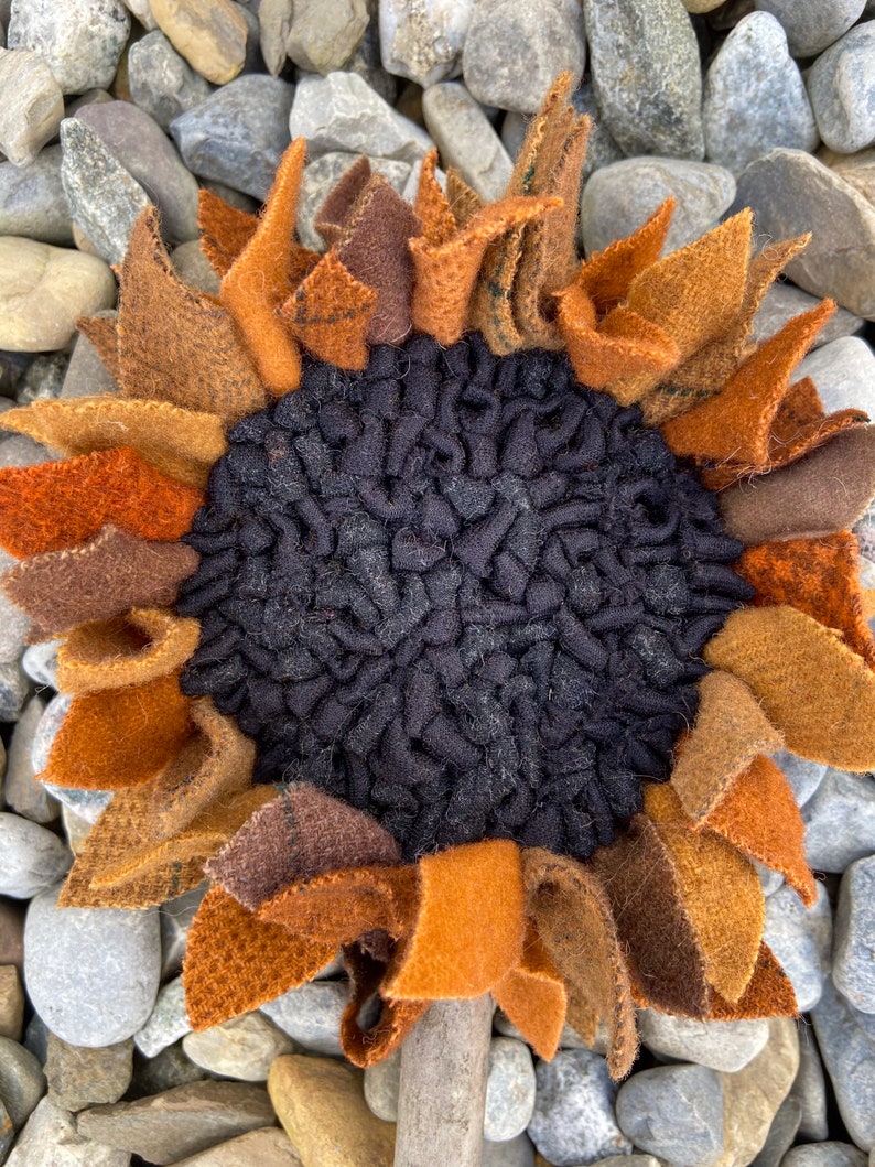 May include: A handmade felt sunflower with brown, orange, and black petals. The sunflower is sitting on a bed of gray rocks.