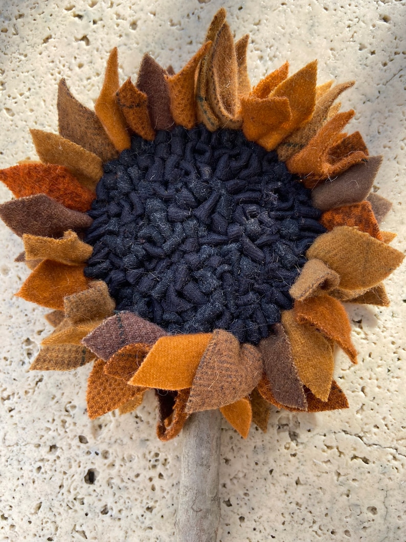 May include: A handmade felt sunflower with a brown, orange, and black color scheme. The sunflower has a textured center and a wooden stem.