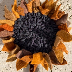 May include: A handmade felt sunflower with brown, orange, and yellow petals. The center of the sunflower is black and textured.