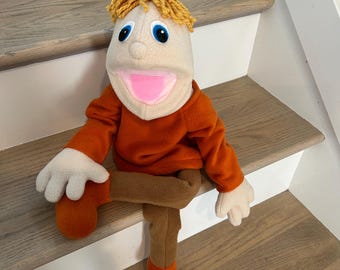 May include: A hand puppet with a beige fabric head, blue eyes, and yellow yarn hair. It wears a long-sleeved orange shirt, brown pants, and orange shoes. The puppet has a wide, open mouth with a pink interior and is posed in a seated position.
