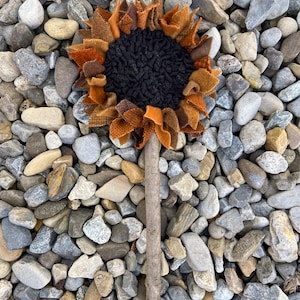 May include: A brown and orange fabric sunflower with a black center, attached to a brown stick. The sunflower is sitting on a bed of gray and brown rocks.