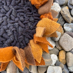 May include: A close-up of a brown, orange, and black felt sunflower with a textured center. The sunflower is sitting on a bed of gray and brown rocks.