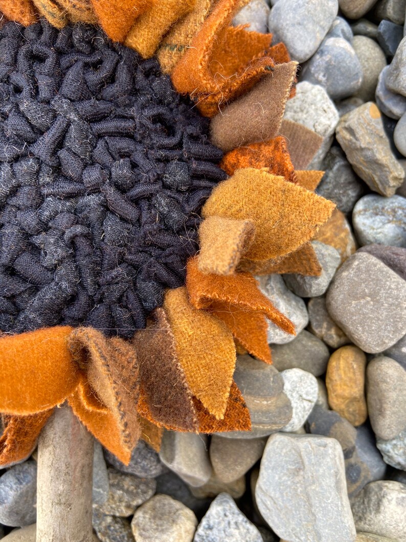 May include: A close-up of a brown, orange, and black felt sunflower with a textured center. The sunflower is sitting on a bed of gray and brown rocks.