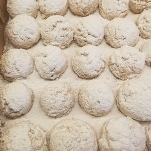 May include: A close-up of a box filled with a large quantity of round, white, powdered sugar cookies. The cookies are arranged in rows and are all the same size and shape.