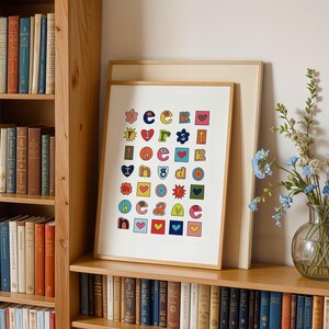 May include: A colourful alphabet print with a white background. The letters are in different colours and shapes, with some letters having hearts, stars, and other designs. The print is framed in a white frame and is hanging on a wall.