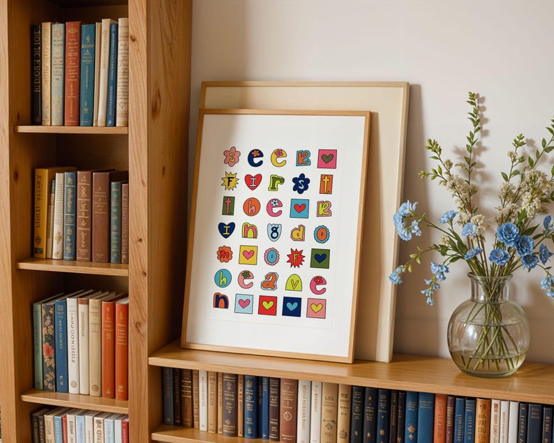 May include: A colourful alphabet print with a white background. The letters are in different colours and shapes, with some letters having hearts, stars, and other designs. The print is framed in a white frame and is hanging on a wall.