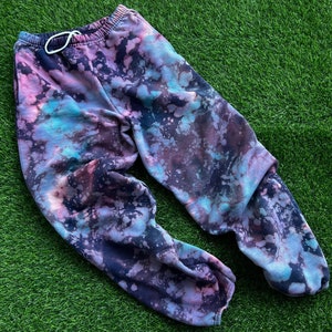 Może przedstawiać: Spodnie dresowe w stylu tie-dye w odcieniach niebieskiego, różowego i czarnego. Spodnie dresowe mają sznurek w pasie i luźny krój.