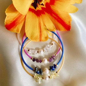 May include: Three stacked bracelets with different coloured bands and gemstone charms. The top bracelet has clear stones, the middle bracelet has purple stones, and the bottom bracelet has blue stones. Each bracelet has a pearl charm.