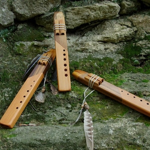 May include: Three wooden flutes with different designs and lengths, resting on a mossy rock surface. The flutes are made of natural wood with a brown and light brown colour scheme.