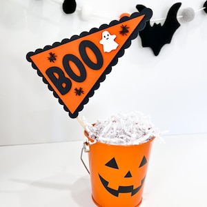 May include: Orange pumpkin-shaped bucket with a black jack-o'-lantern face. The bucket is filled with white shredded paper and a black and orange pennant flag with the word "BOO" in black letters and a white ghost.